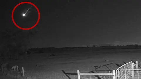 A meteor, circled with a red overlay, streaks over a field at night. A fence is pictured in the foreground of the image and a thin line of trees is faintly visible in the distance.