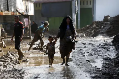 woman holds child by the hand while walking barefoot on a street covered with mud