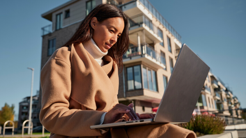 A young woman is sitting outdoors in a modern residential neighbourhood, concentrating on her laptop. She is wearing a beige coat and a white turtleneck jumper. Modern flats with balconies can be seen in the background.