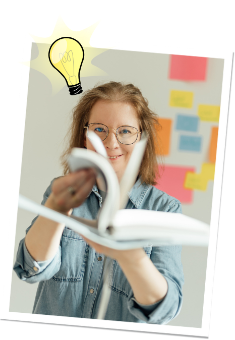 Das hochgeladene Bild zeigt eine Frau mit Brille, die ein aufgeschlagenes Buch in der Hand hält und in die Kamera lächelt. Sie trägt ein Jeanshemd, und im Hintergrund sind bunte Haftnotizen an der Wand angebracht. Oben links ist eine gezeichnete Glühbirne zu sehen, die eine Idee oder Inspiration symbolisiert. Das Bild vermittelt Kreativität und Wissensaustausch.