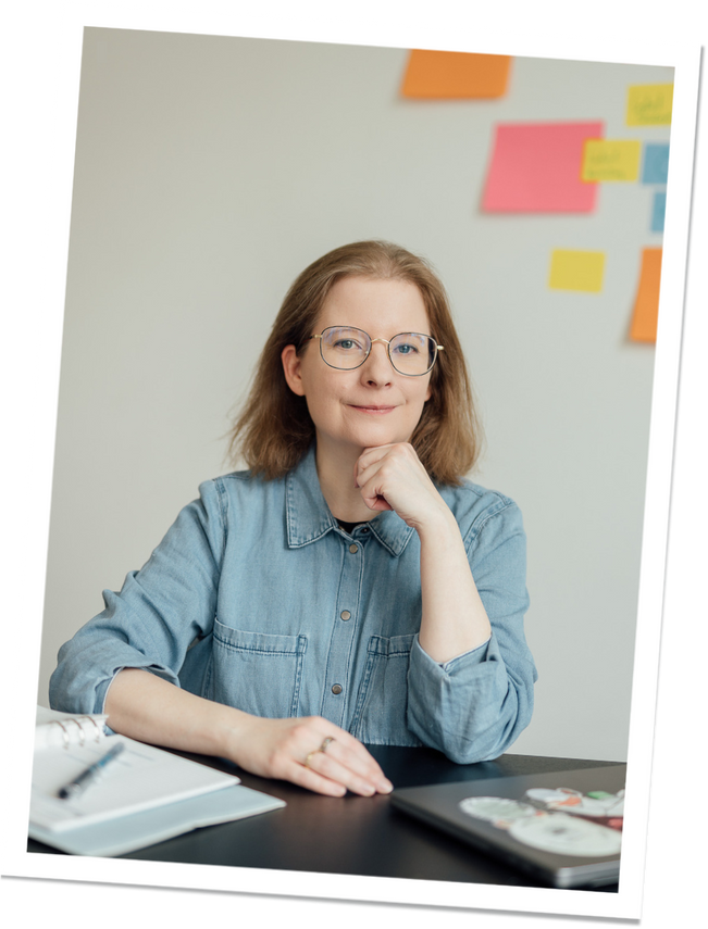 Eine Frau mit schulterlangem, hellbraunem Haar sitzt an einem Schreibtisch. Sie trägt eine Brille und ein Jeanshemd. Im Hintergrund sind bunte Haftnotizen an der Wand angebracht.