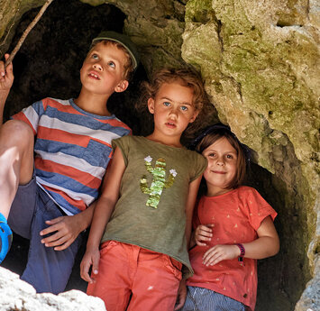 In einer Felswand ist ein großes Loch in dem zwei Mädchen und ein Junge mit einem Stock in der Hand stehen.